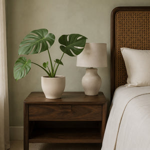 Monstera artificielle 60 cm sur une table de chevet en bois, à côté d'une lampe éteinte